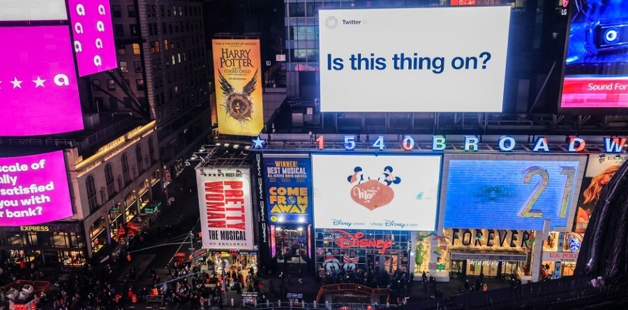 A busy New York City street with various billboards around. One looks like a giant Tweet with the words "Is this thing on?"