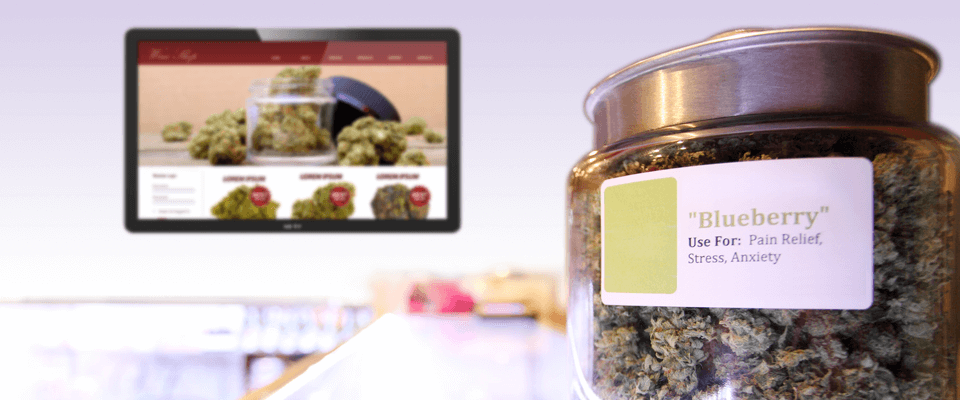 A glass jar filled with cannabis. In the background, digital signage is delivering information to individuals inside of the dispensary
