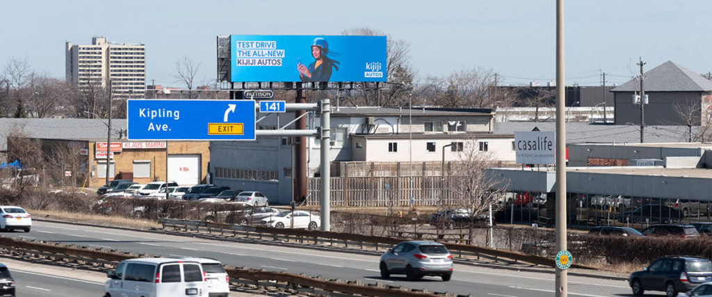 A busy roadway. Above and in the background, a Pattison digital billboard displays an ad.