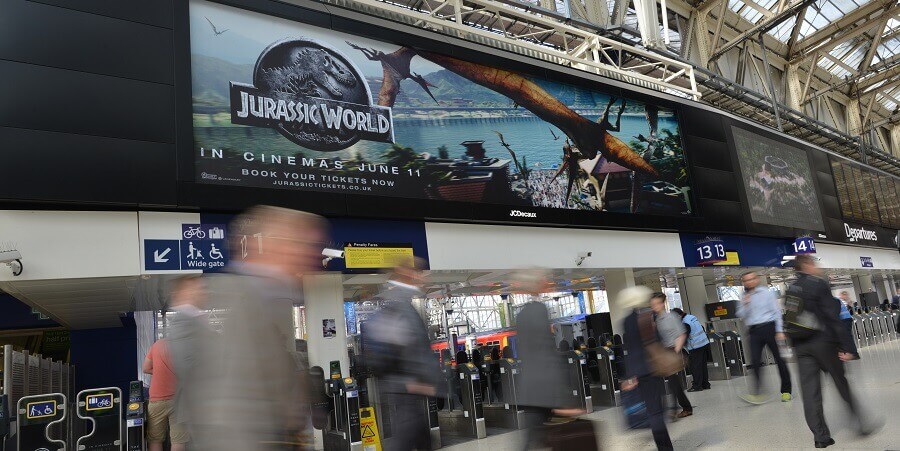 Transit users walking through Waterloo station. Overhead, a large digital billboard displays an ad for the film "Jurassic World." Transit hubs are high-traffic areas perfect for connecting people with favourite brands.