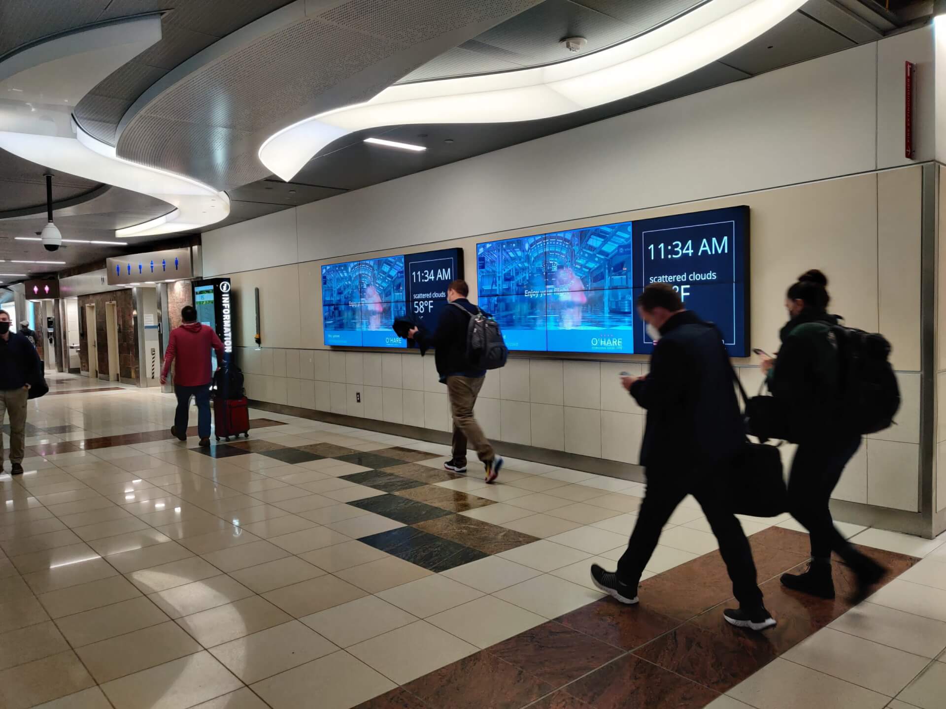 Travelers walking through an airport concourse past large digital out-of-home advertising screens displaying weather information and airport messaging.