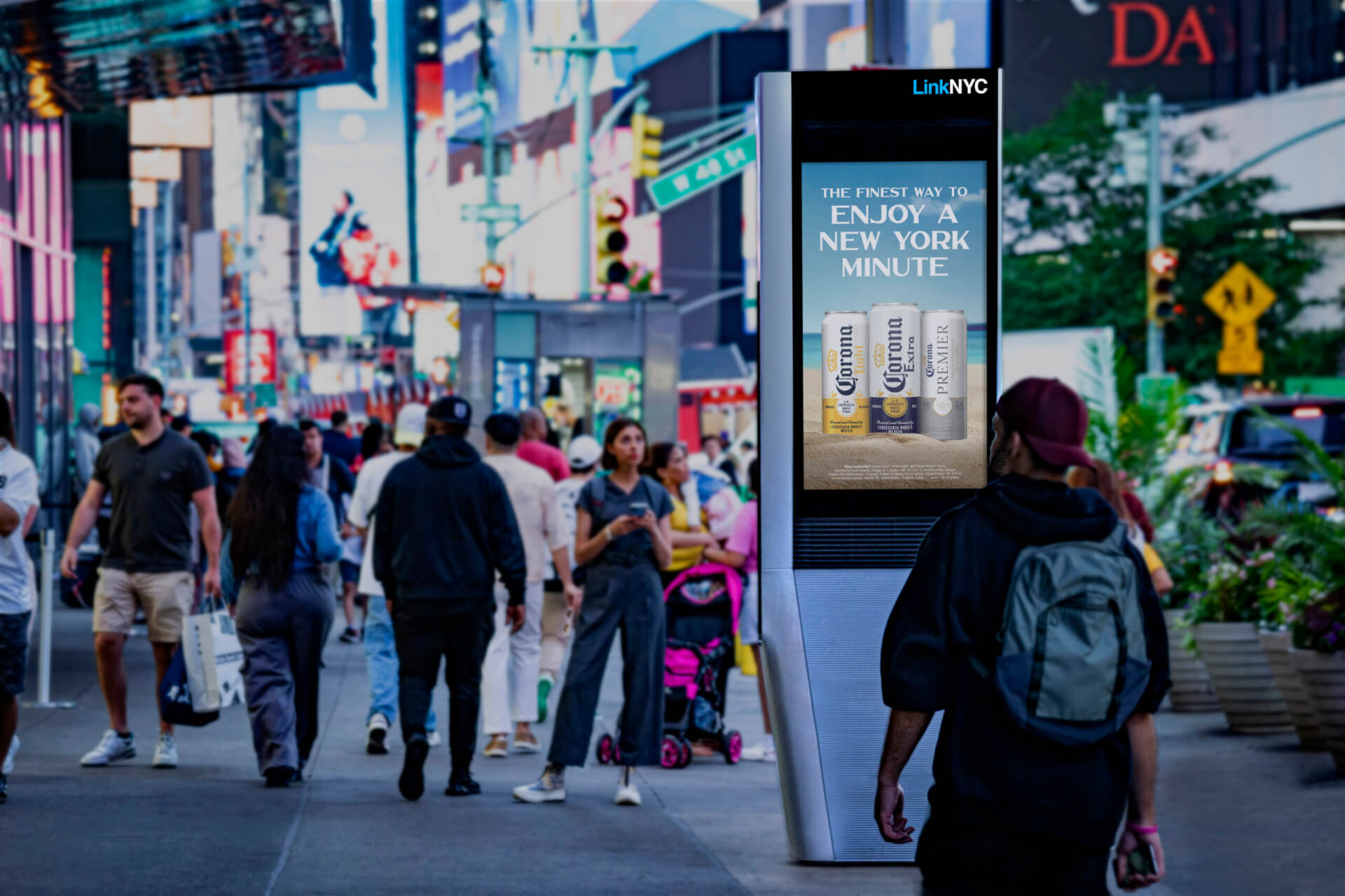 A busy New York City sidewalk with pedestrians walking past a LinkNYC digital kiosk displaying a Corona beer advertisement. The kiosk stands prominently in the foreground against a backdrop of bright city lights, storefronts, and traffic, illustrating digital out-of-home advertising in a high-footfall urban environment.