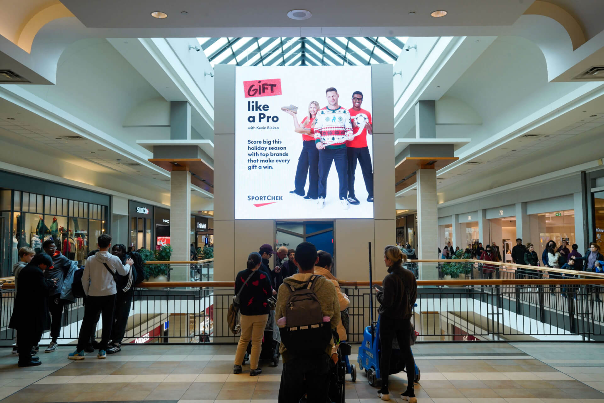 A busy two-level shopping mall atrium with shoppers gathered along the upper-floor railing. A large digital billboard dominates the center, displaying a SportChek holiday advertisement beneath a glass skylight.