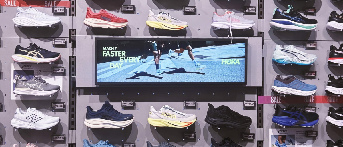 In-store shoe display featuring running footwear from HOKA, ASICS, New Balance and other brands, with a digital screen showing a HOKA advertising campaign.